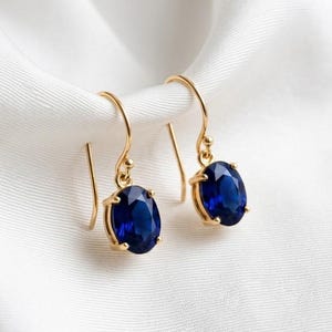 May include: A pair of gold drop earrings featuring oval-shaped deep blue gemstones. Each earring is suspended from a curved gold wire hook, with the blue stone held in a four-prong setting. The design is simple, focusing on the contrast between the gold metal and the vibrant blue colour of the stones.