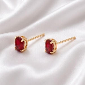 Puede incluir: Un par de pendientes de botón dorados con piedras preciosas de color rojo intenso en forma ovalada, sujetas por engastes de cuatro garras. Los pendientes tienen postes finos y rectos que salen de la parte posterior de la montura.