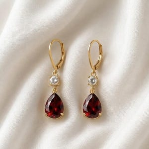 Può includere: Un paio di orecchini in oro con chiusura a monachella, caratterizzati da una piccola pietra rotonda trasparente sopra una pietra granato rosso scuro a forma di goccia. La montatura in metallo sostiene le pietre sfaccettate in posizione.