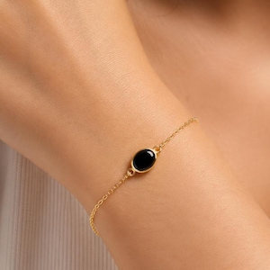 May include: A delicate gold chain bracelet featuring a single, smooth, oval-shaped black stone set in a gold bezel. The bracelet is secured with a thin, polished gold link chain that wraps around the wrist, providing a simple and refined appearance.