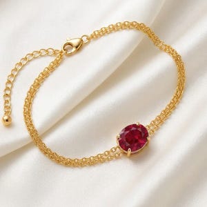 May include: A gold-toned bracelet featuring a double chain design and a single oval-shaped deep red gemstone set in a prong setting. The bracelet includes a lobster claw clasp and an adjustable extender chain with a small round bead at the end.