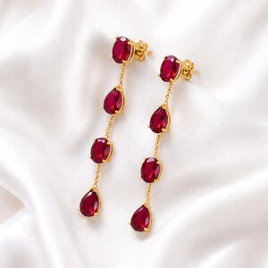 Puede incluir: Un par de pendientes largos de oro con cuatro piedras facetadas de color rojo intenso, de formas ovalada y de pera, suspendidas en línea vertical. Las piedras están engastadas en metal dorado y unidas por delicadas cadenas de oro.