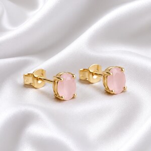 Puede incluir: Un par de pendientes de botón con piedras de color rosa claro en forma ovalada engastadas en metal dorado pulido. Cada pendiente tiene un cierre de poste seguro y un engaste de garras que sujeta el cabujón rosa liso y translúcido.