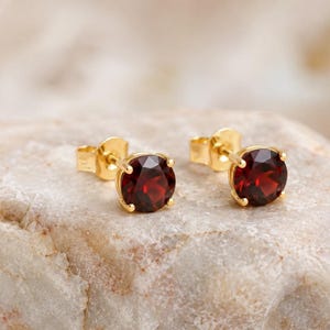Puede incluir: Un par de pendientes de botón con piedras preciosas de granate rojo oscuro redondas engastadas en oro amarillo pulido. Cada piedra está asegurada por un engaste de cuatro garras, mostrando una superficie facetada que refleja la luz.