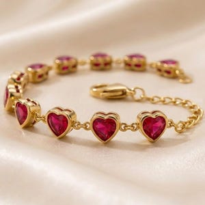 May include: A gold-toned chain bracelet featuring a series of vibrant, deep red heart-shaped stones set in metal frames. The bracelet includes a lobster claw clasp and an adjustable chain link extension for a secure fit around the wrist.