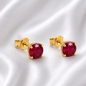 May include: A pair of stud earrings featuring round, faceted, deep red stones set in polished yellow gold. Each earring has a classic four-prong setting and a secure post back, showcasing a brilliant cut that reflects light across the surface.