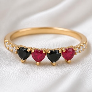 Puede incluir: Un anillo de banda dorada con un patrón alterno de dos piedras negras en forma de corazón y dos piedras rojas en forma de corazón en el centro, flanqueadas por pequeñas piedras redondas transparentes engastadas a lo largo de la banda de metal pulido.