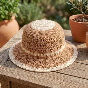 May include: A crochet bucket hat featuring a rounded crown and a wide brim. The hat is crafted with alternating bands of tan and cream-colored yarn, with a decorative scalloped edge detail in cream around the outer circumference of the brim.