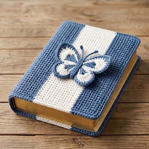Può includere: Una copertina per libro rettangolare realizzata in filato lavorato all'uncinetto con strisce verticali blu polvere e bianco. Una farfalla tridimensionale all'uncinetto in blu e bianco coordinato è fissata al centro. Il libro ha bordi dorati.