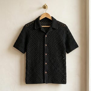 May include: A black short-sleeved shirt crafted with a crochet granny square pattern. The shirt features a classic collar, a button-down front with five brown buttons, and a semi-sheer open-knit texture throughout the entire garment.