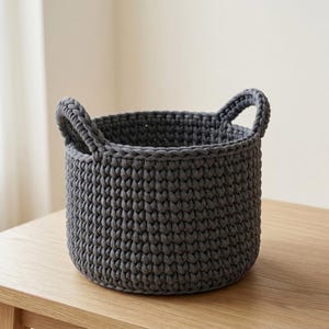 May include: A dark gray round storage basket crafted from thick, chunky crocheted yarn. The basket features a deep, textured knit pattern and two integrated handles on opposite sides, providing a sturdy structure for holding various household items.