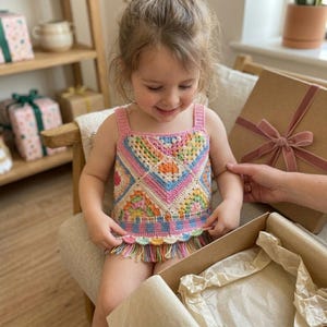 May include: A sleeveless crochet top featuring a multi-colored granny square pattern in shades of pink, yellow, blue, and orange. The garment has a square neckline, a checkered band at the waist, and a scalloped hem finished with long, multicolored fringe.