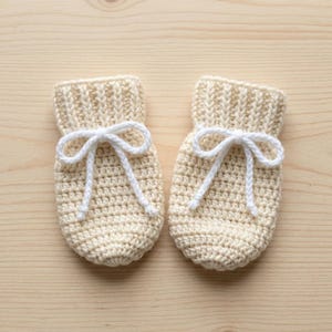 May include: A pair of cream-colored crochet baby mittens featuring a ribbed cuff and a white braided drawstring tie at the center. The mittens are constructed with a dense stitch pattern and have a rounded shape at the bottom for a snug fit.