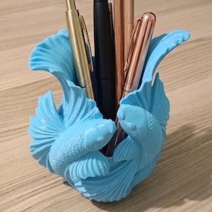 Könnte beinhalten: Ein hellblauer Stiftehalter in Form von zwei Kampffischen mit fließenden Flossen. Der Halter enthält einen goldenen Stift, einen schwarzen Stift, einen Holzbleistift mit blauem Schmucksteinaufsatz und einen roségoldenen Stift.