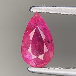 Puede incluir: Una piedra preciosa facetada en forma de pera con un color rosa intenso y vibrante. La piedra presenta un corte claro y simétrico con múltiples facetas triangulares y en forma de cometa que reflejan la luz, sujeta por unas pinzas metálicas dentadas.