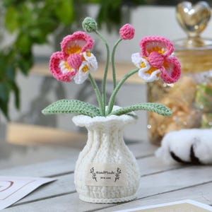 Puede incluir: Un jarrón blanco de ganchillo con un diseño texturizado y acanalado, con una etiqueta rectangular que dice HandMade WITH LOVE. Contiene dos flores de ganchillo con pétalos rosas, blancos y amarillos, tallos verdes y dos pequeños capullos con hojas.