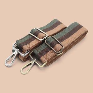Pode incluir: Duas alças ajustáveis para bolsa com um padrão de listras tecidas em tons de verde oliva, castanho escuro e bege. Cada alça inclui uma fivela de metal deslizante para ajuste de comprimento e um gancho mosquetão giratório em prata e bronze.