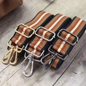 Pode incluir: Três alças ajustáveis para bolsa com padrão de listras em tons de marrom, branco e preto. Cada alça inclui uma fivela deslizante e dois mosquetões giratórios de metal. Os acabamentos das ferragens incluem tons de dourado, prateado e chumbo.