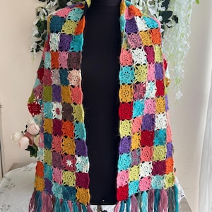 Puede incluir: Un chal de ganchillo hecho a mano con un vibrante mosaico de cuadrados de la abuela multicolores en tonos rojo, amarillo, azul, verde, morado y rosa. La prenda rectangular está rematada con largos flecos de hilo en el borde inferior.