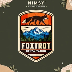May include: A shield-shaped sticker featuring a black fox silhouette against an orange sky, above a blue mountain range and green pine forest. The text FOXTROT and DELTA TANGO is printed in white on a black background within the sticker design.