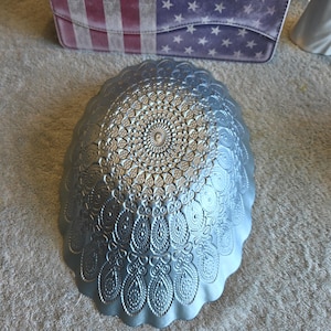 May include: A silver-colored oval bowl featuring a detailed, embossed mandala-style pattern across its entire surface. The bowl has a scalloped edge and a metallic finish, displaying intricate geometric and teardrop-shaped textures throughout.