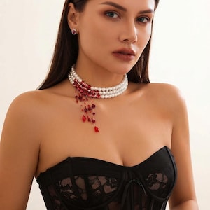Puede incluir: Un collar tipo gargantilla de tres hileras de perlas con un degradado rojo en un lado, adornado con cuentas de cristal rojas colgantes y pequeños detalles florales rojos. Se lleva con un corsé de encaje negro y un pendiente de botón rojo.