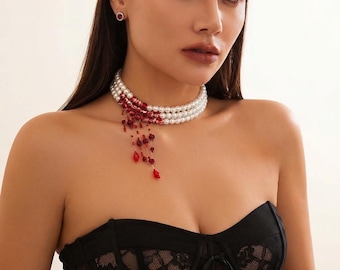 May include: A three-strand pearl choker necklace featuring a red gradient effect on one side, with dangling red crystal beads and small red floral accents. The necklace is worn with a black lace corset top and a single small red stud earring.