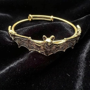Op de afbeelding: Een goudkleurige metalen armband met een centraal vleermuismotief met gedetailleerde vleugels die zich langs de band uitstrekken. De vleermuis is in het midden geplaatst, met de vleugels wijd gespreid, wat een doorlopend decoratief element vormt.