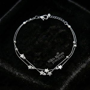 Pode incluir: Uma pulseira de tom prateado com fio duplo, apresentando pequenos berloques de estrelas metálicas e contas redondas intercaladas ao longo das correntes delicadas. A peça inclui um fecho lagosta e uma corrente extensora. O texto Nikis Bloom Jewellery é visível.