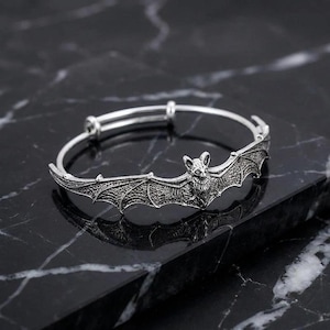 Op de afbeelding: Een zilverkleurige metalen armband met een centraal vleermuismotief, gedetailleerde vleugels en textuur in de vacht. De armband heeft een verstelbaar open ontwerp met een schuifmechanisme en een geoxideerde afwerking die de gravures benadrukt.