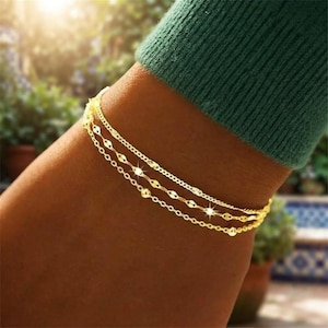 Puede incluir: Una pulsera de cadena de tres hilos en tono dorado con eslabones delicados y pequeñas cuentas esféricas. La pulsera se lleva en la muñeca, parcialmente cubierta por una prenda de manga larga de color verde oscuro, con un fondo exterior soleado.