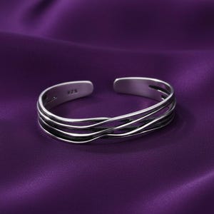 Könnte beinhalten: Ein offener Ring in Silberoptik mit einem mehrschichtigen, wellenförmigen Design aus drei Bändern, die sich überlappen und ineinander verschlingen. Die Innenseite des Metallbandes ist deutlich mit dem Stempel 925 für Sterlingsilber versehen.