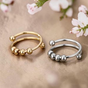 May include: Two open-ended rings, one in a gold tone and one in a silver tone, each featuring a thin band with four small, movable spherical beads attached to the front. The rings are positioned side by side, showcasing their metallic finish.