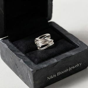 Op de afbeelding: Een zilveren ring met meerdere overlappende, dunne draadbanden in een chaotisch, gelaagd ontwerp. De ring wordt gepresenteerd in een zwart, gestructureerd juwelendoosje met de tekst Nikis Bloom Jewelry gedrukt op de voorste hoek.
