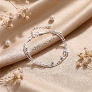 Puede incluir: Una pulsera de plata de varias hebras con cadenas delicadas y pequeñas cuentas de plata pulida distribuidas a lo largo de las hebras. La pulsera incluye un cierre de mosquetón y una cadena extensora. El texto NikisBloomJewellery es visible.