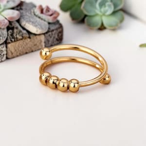 May include: A gold-toned metal ring featuring a bypass design with an open band. The ring includes four small, smooth, spherical beads that are positioned along the band and can slide freely, creating a functional and polished jewelry piece.
