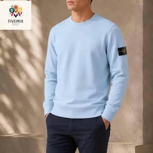 May include: A light blue long-sleeve crewneck sweatshirt featuring a ribbed collar, cuffs, and hem. A black and yellow circular patch logo is attached to the upper left sleeve. The garment is paired with dark navy blue trousers.
