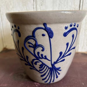 May include: A light gray stoneware crock featuring a hand-painted cobalt blue bird design. The vessel has a wide, flared rim and a tapered body. The blue pattern depicts a stylized bird with intricate, flowing lines and decorative leaf-like accents.