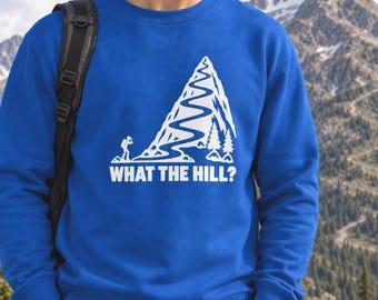 Peut inclure: Un sweat-shirt bleu roi à col rond arborant un motif blanc représentant un sentier sinueux sur une montagne, un randonneur avec un sac à dos et deux sapins. Sous le motif, le texte "WHAT THE HILL?" est écrit en lettres blanches grasses.