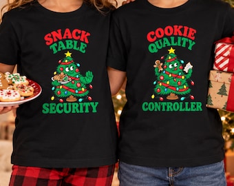 Peut inclure: Deux t-shirts noirs avec un motif de sapin de Noël animé. Le t-shirt de gauche porte l'inscription SNACK TABLE SECURITY avec un sapin tenant une assiette de biscuits. Celui de droite indique COOKIE QUALITY CONTROLLER avec un bonhomme en pain d'épice.