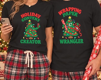 Peut inclure: Deux t-shirts noirs avec des graphismes de sapin de Noël. Le t-shirt de gauche montre un sapin prenant un selfie avec le texte HOLIDAY CONTENT CREATOR. Celui de droite montre un sapin emmêlé dans du ruban avec le texte WRAPPING PAPER WRANGLER.