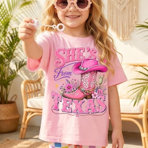 May include: A light pink t-shirt featuring a graphic of a pink cowboy boot with a matching hat, surrounded by flowers and a cactus, with the text "She's From TEXAS" in pink lettering. The shirt is worn with a lavender headband and flower-shaped sunglasses.