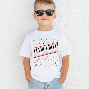 May include: A white short-sleeved t-shirt featuring a graphic print of a diagonal piano keyboard section bordered by two red lines, surrounded by scattered black musical notes and symbols across the front of the garment.