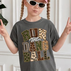 May include: A charcoal gray short-sleeve t-shirt featuring the text RUN THE DANG BALL in large block letters. Each letter contains various football-themed patterns including footballs, field markings, helmets, and play diagrams in green, brown, and white.