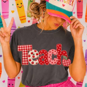 Op de afbeelding: Een antracietgrijs t-shirt met het woord TEACH in grote, bedrukte stoffen letters met rood gingham, appelprints, rode bloemen en roze hartjes. Een witte en blauwgroene truckerpet met het woord TEACHER in regenboogkleurige letters.