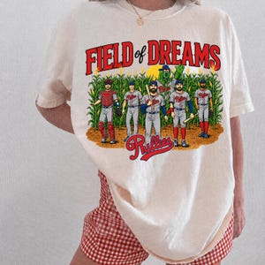 May include: A cream-colored t-shirt featuring a graphic print with the text FIELD OF DREAMS in red. The illustration shows five baseball players in Phillies uniforms and a green mascot standing in a cornfield, with the Phillies logo below.