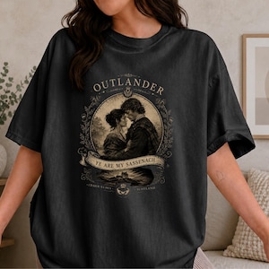Könnte beinhalten: Ein anthrazitfarbenes Kurzarm-T-Shirt mit einem kreisförmigen Grafikdruck eines Paares in historischer Kleidung. Der Text lautet OUTLANDER, YE ARE MY SASSENACH, CRAIGH NA DUN und SCOTLAND, umgeben von einem verzierten botanischen Rahmen.