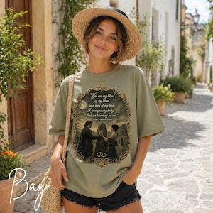 May include: A sage green t-shirt featuring a graphic print of a couple standing before a stone monolith within an ornate gold frame. The text reads: You are my blood of my blood, and bone of my bone. I give you my body, that we two may be one.