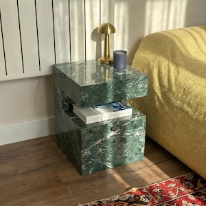 May include: A square, dark green marble side table with a C-shaped profile featuring a hollow middle section. A white book titled Simplicity rests in the center opening. A gold-toned mushroom-shaped lamp and a gray candle sit on the top surface.