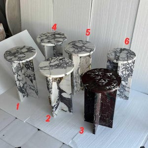 May include: Six cylindrical side tables made of polished marble, each featuring a cross-shaped base. Five tables display white marble with black veining, while one table is crafted from deep red marble with white veining. Each table is labeled with a red number.